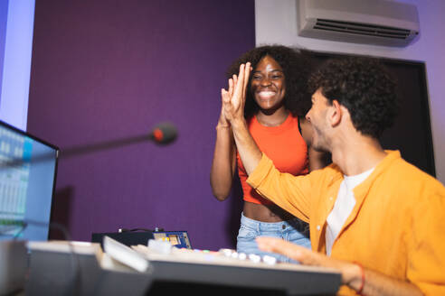 Positive male audio engineer showing like gesture to excited female African American vocalist in control room while recording song in musical studio - ADSF47502