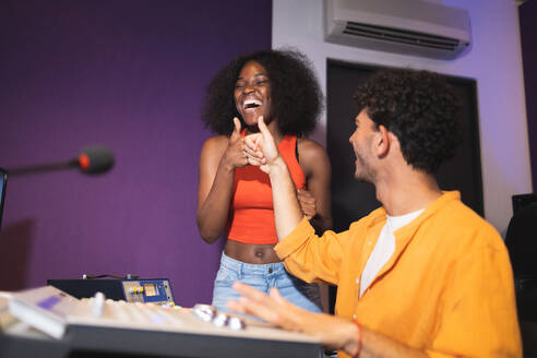 Positive male audio engineer showing like gesture to excited female African American vocalist in control room while recording song in musical studio - ADSF47503