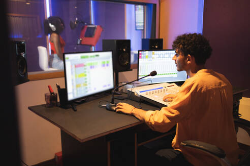 Side view of male audio engineer using mixing board and computer sound software while African American female performer singing song in musical studio - ADSF47506
