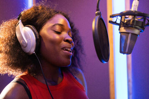 Side view of African American female professional vocalist in headphones taking high note near microphone while singing and recording in musical studio - ADSF47508