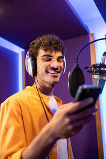 Male professional singer in headphones near microphone listening to recorded vocal sound on cellphone while working in production musical studio - ADSF47510