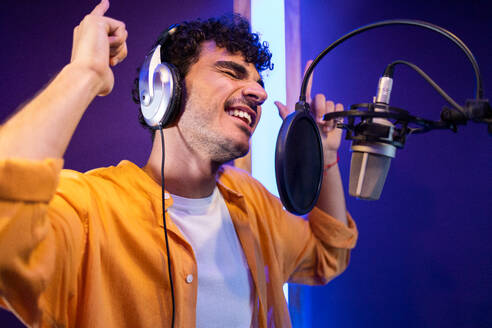 Side view of male professional singer in headphones taking high note near microphone and pointing up while creating music in studio - ADSF47511