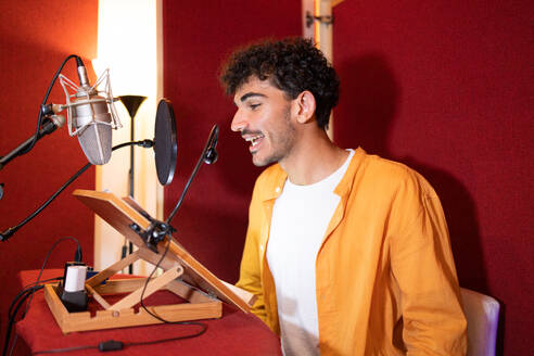 Side view of smiling young Hispanic male radio host looking away while sitting at desk with microphone equipment and recording podcast in studio room - ADSF47516