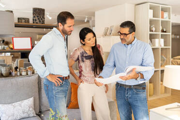 Focused positive couple clients in casual clothes standing and checking furniture catalogue on table with mature male attendant in furniture store - ADSF47522