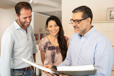 Focused crop positive couple clients in casual clothes standing and checking furniture catalogue on table with mature male attendant in furniture store - ADSF47523