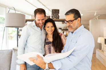 Focused positive couple clients in casual clothes standing and checking furniture catalogue on table with mature male attendant in furniture store - ADSF47524