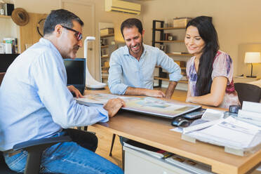 Happy confident couple sitting at table with manager of furniture store while looking at catalogue and selecting furniture products in modern office - ADSF47527