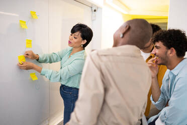 Side view of female manager looking down while standing and sticking yellow notes on white board in front of young multiracial team members in room - ADSF47551