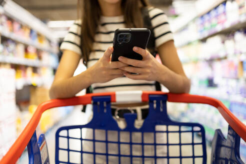 Crop anonymous female customer in casual clothes using smartphone while shopping alone at supermarket - ADSF47561