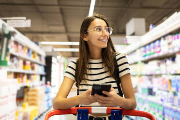 Positive young female customer in casual clothes and eyeglasses using smartphone while shopping alone at supermarket - ADSF47562