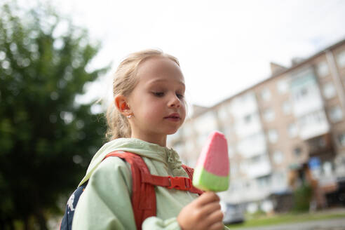 Side view from below of a child with a backpack, standing on the grass and holding an ice cream in his hand in a faded housing complex - ADSF47564