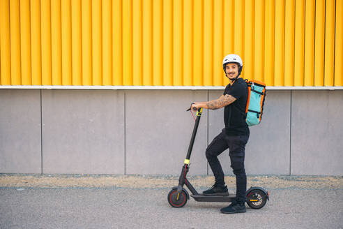 Full body side view of happy delivery man in helmet standing on electric kick scooter on asphalt road near building wall in city - ADSF47571