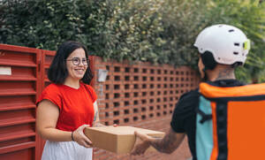 Cheerful young female in casual clothes receiving package from anonymous delivery man in protective helmet on sidewalk - ADSF47575