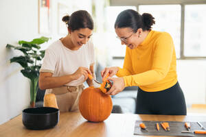 Smiling young Hispanic female friends looking down while standing at table with Halloween tools and pulling out pulp from top of orange pumpkin in daylight - ADSF47576