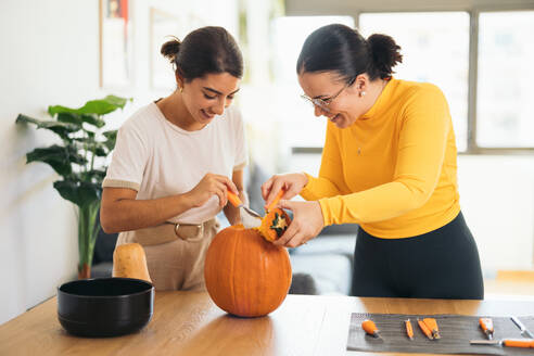 Smiling young Hispanic female friends looking down while standing at table with Halloween tools and pulling out pulp from top of orange pumpkin in daylight - ADSF47576