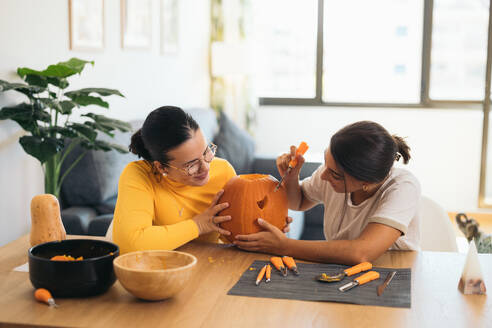 Focused young Hispanic female friends looking down while sitting at table with Halloween kit bowls and carving eye with sharp tool on orange pumpkin - ADSF47579