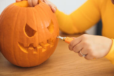 Anonymous female sitting at table and carving with tool scary teeth on orange pumpkin and preparing for Halloween decoration in day - ADSF47581