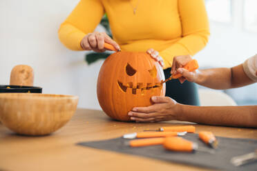 Anonymous women near table carving with tool scary teeth on orange pumpkin and preparing for Halloween decoration in day - ADSF47582