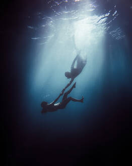 Side view of unrecognizable couple in swim cloth holding hands while diving together in wetsuit and snorkeling mask under blue sea at night with bright light - ADSF47604