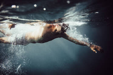 Underwater view of unrecognizable crop man in swimming goggle diving in blue sea water with bubbles in sunlight - ADSF47605
