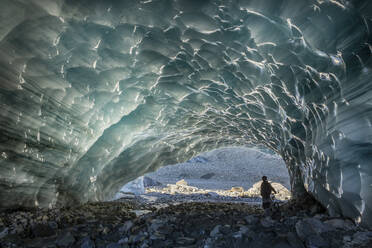 Back view of anonymous tourist standing near bike under hole while exploring ice cave in Switzerland on winter day - ADSF47615