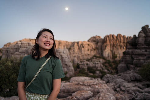 Fröhliche asiatische Reisende, die wegschaut und die herrliche Aussicht auf die Sierra del Torcal in Antequera genießt, während sie sich bei Sonnenuntergang vor dem Mond entspannt - ADSF47631