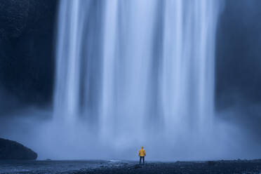 Back view of anonymous tourist in raincoat standing in front of large waterfall with clean water during trip - ADSF47632