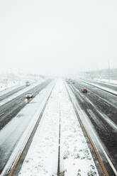 Cars driving on smooth asphalt roadway covered with snow in gloomy winter day during snowfall in Madrid - ADSF47654