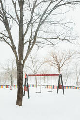 Playground with swings and slide covered with snow among bare trees in winter day in gloomy weather in Madrid - ADSF47655