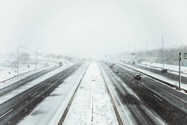 Cars driving on smooth asphalt roadway covered with snow in gloomy winter day during snowfall in Madrid - ADSF47662