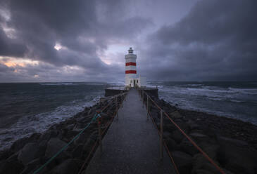 Scenic view of lighthouse with beacon on top located on seashore close to wavy and foamy seawater against dark overcast clouds of sunset time in daylight - ADSF47667