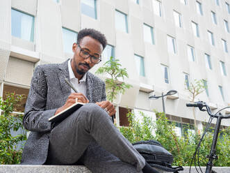 From below African American male with eyeglasses in formal suit sitting on city street and writing notes in notebook - ADSF47672