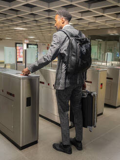 Back view of African American male pedestrian in formal suit with backpack carrying suitcase passing tourniquet with card in underground subway - ADSF47674