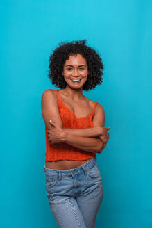 Friendly African American female fashion model in casual clothes with curly hair smiling with crossed arms while standing against blue background - ADSF47687
