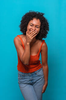 Excited African American female model with Afro hairstyle in casual outfit laughing at joke with closed eyes and covering face against blue background - ADSF47688