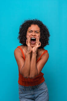Joyful African American female model with Afro hairstyle wearing casual clothes shouting against blue background - ADSF47689