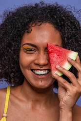 Human face of friendly charismatic African American female model smiling and looking at camera while hiding eye behind slice of sweet watermelon - ADSF47691