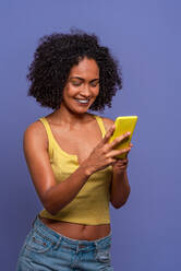 Cheerful Hispanic female with Afro hairstyle in yellow top browsing smartphone while standing on blue background - ADSF47692