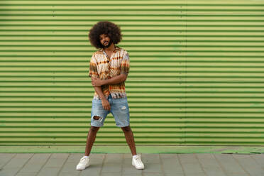 Full body side view of young African American male with curly hair in casual clothes standing along striped green wall - ADSF47699