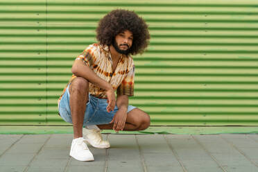 Full body of confident young African American male in trendy clothes with Afro hairstyle looking at camera crouching against green striped wall - ADSF47701