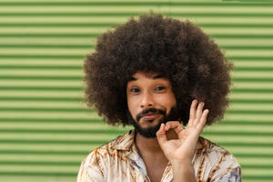 Portrait of smiling African American male in casual clothes with Afro hair making okay gesture and looking at camera against green wall - ADSF47702