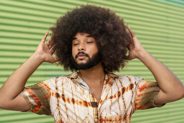 Hispanic man with Afro hairstyle eyes closed and raising arms while pretending to touch curly hair against green ribbed background - ADSF47703