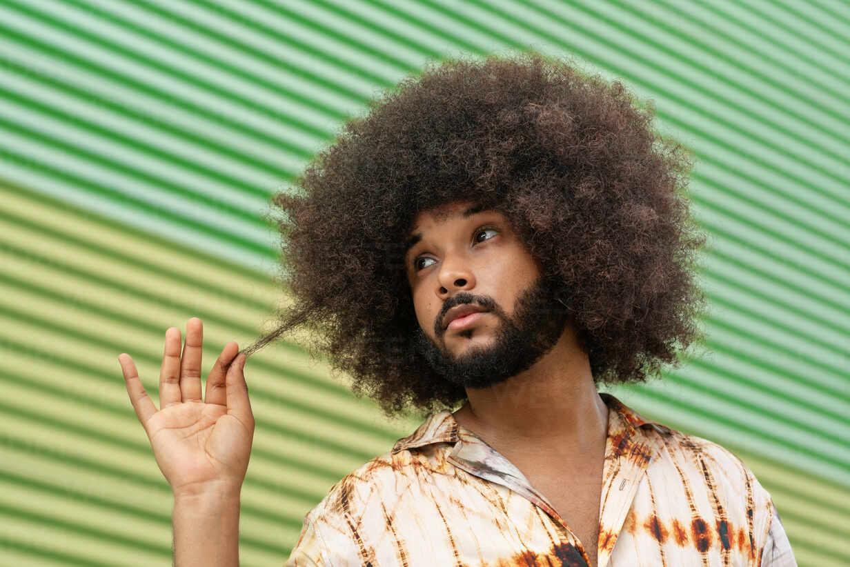 Hispanic bearded male with Afro hairstyle in Hawaiian shirt looking away  thoughtfully while touching hair against ribbed green backdrop stock photo