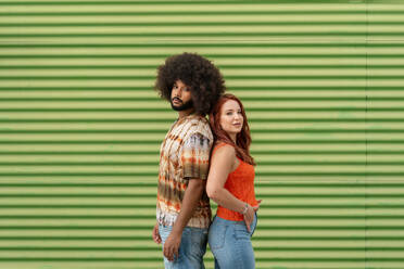 Lady with red hair looking at camera while standing back to back with curly Hispanic male soulmate against green wall - ADSF47709