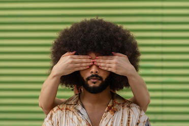 Unrecognizable partner covering eyes of Hispanic bearded male with Afro hairstyle against blurred green ribbed wall - ADSF47710