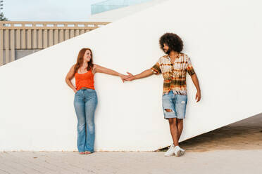 Full length of young African American man wearing summer shirt and denim shorts holding hands with redhead girlfriend while next to a white wall - ADSF47711