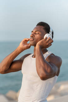 Side view of pensive African American male with closed eyes standing near sea and listening to music in wireless headphones - ADSF47712