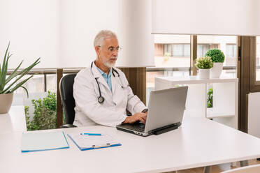 Senior male practitioner in medical uniform with stethoscope chatting with patient while working at desk with netbook and clipboard in clinic - ADSF47715