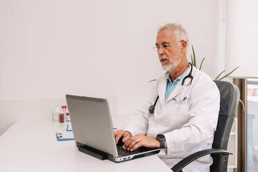 Senior male practitioner in medical uniform with stethoscope chatting with patient while working at desk with netbook and clipboard in clinic - ADSF47716