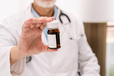 Unrecognizable bearded male doctor in medical uniform with stethoscope showing bottle of medicine in hospital - ADSF47718